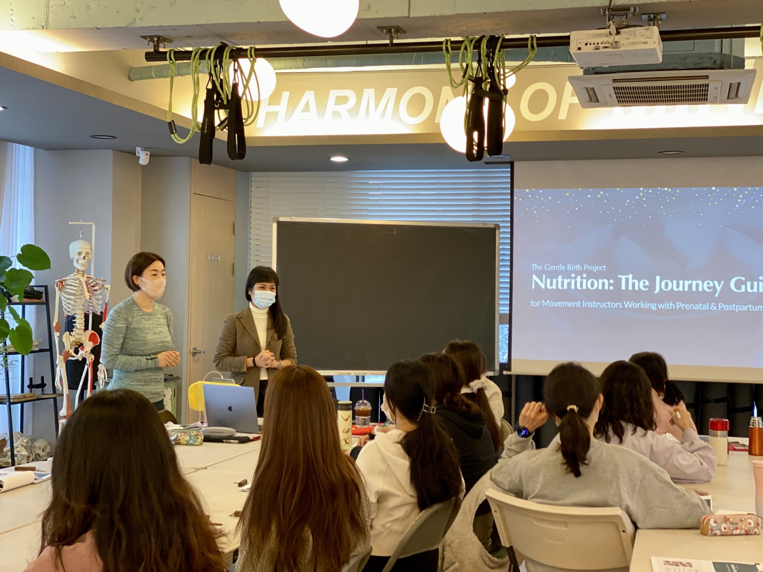 A group of people seated at tables watch two presenters, one masked, discussing a nutrition guide for peak performance. The presentation title "Nutrition: The Journey Guide" is displayed on a screen.