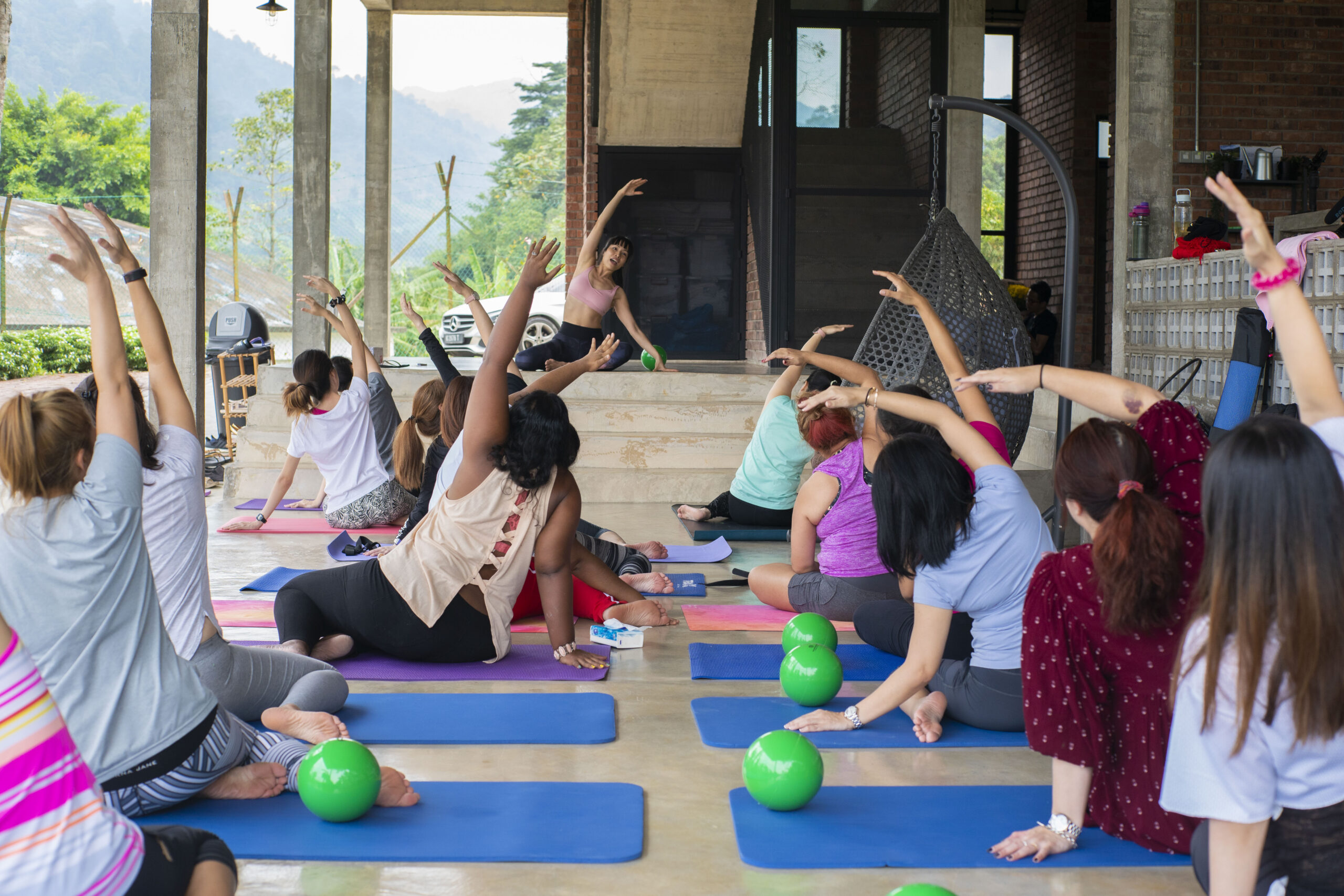 A group of people are participating in a yoga class outdoors, practicing stress management techniques while seated on colorful mats. Green exercise balls are placed nearby on the floor.